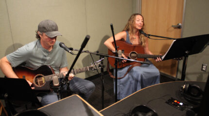 Two adults playing acoustic guitar in a radio studio, one singing into a microphone
