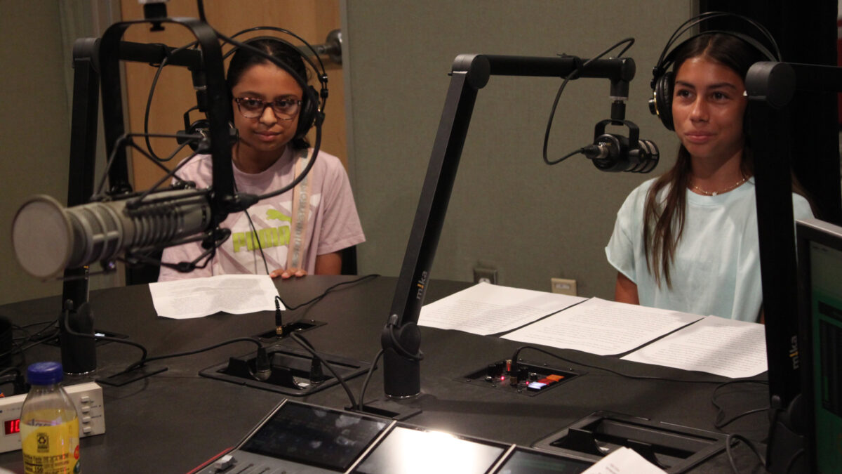 Two teens wearing headphones sitting behind microphones and pieces of paper in a radio studio