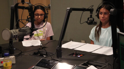 Two teens wearing headphones sitting behind microphones and pieces of paper in a radio studio