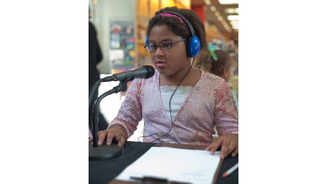 A youth wearing headphones sitting behind a clipboard and speaking into a microphone