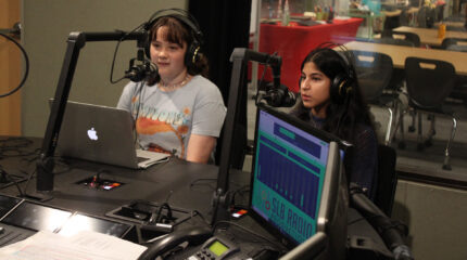 Two teens wearing headphones speaking into microphones in a radio studio