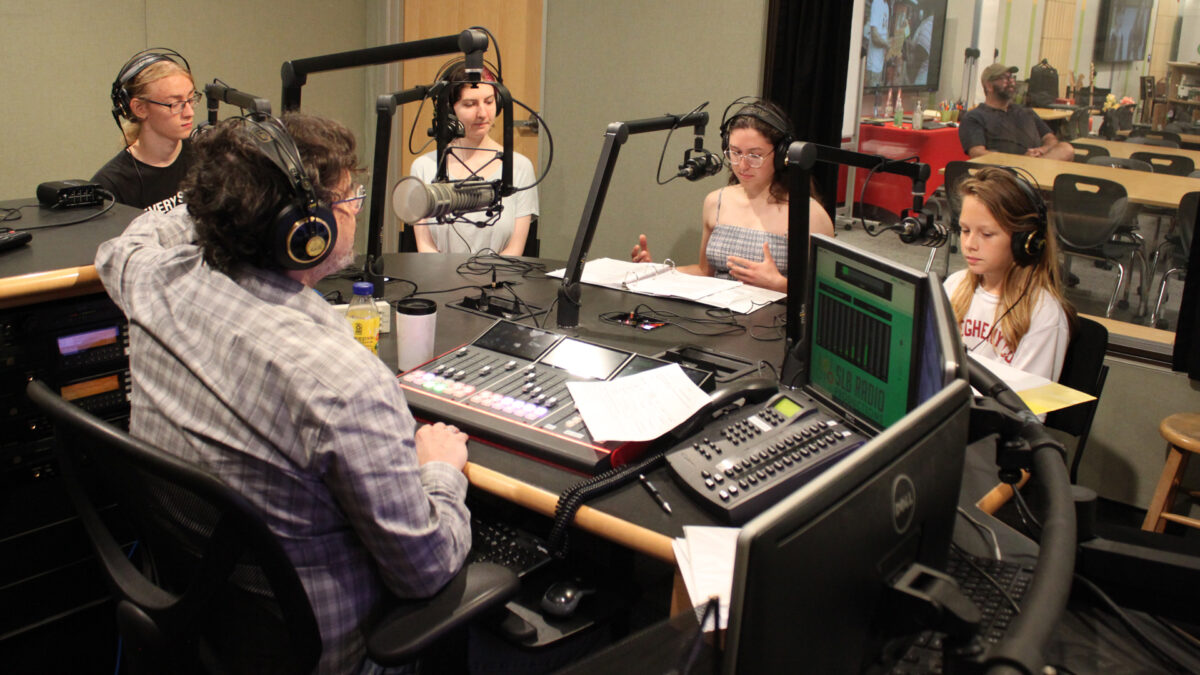 Four teens wearing headphones sitting behind microphones being interviewed by an adult wearing headphones sitting behind a microphone and an audio mixer in a radio studio