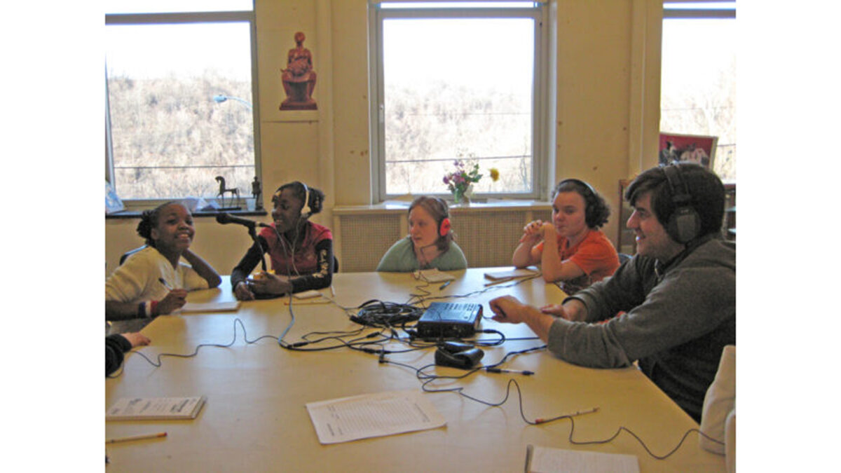 Four youth wearing headphones and an adult wearing headphones sitting around a microphone and a portable audio mixer in a classroom