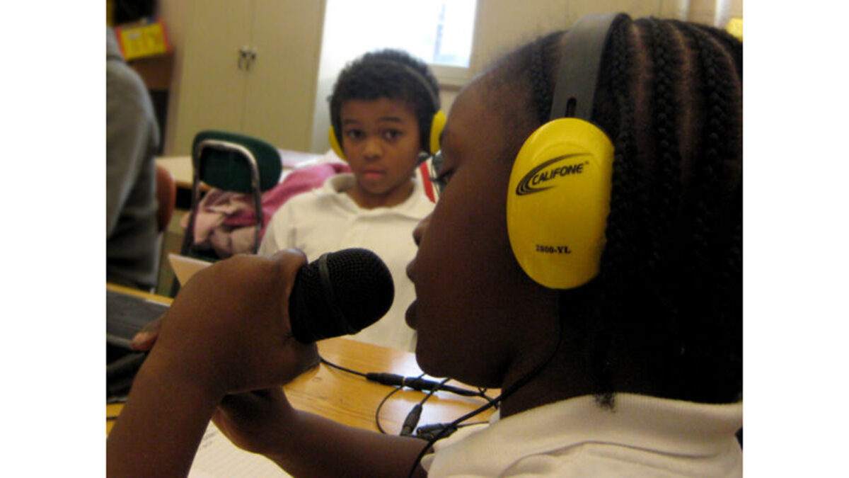 Two youth wearing headphones in a classroom, one holding and speaking into a microphone