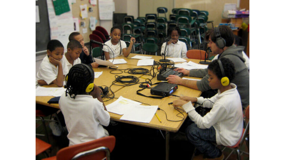 Eight youth wearing headphones and an adult sitting around microphones and a portable audio mixer in a classroom