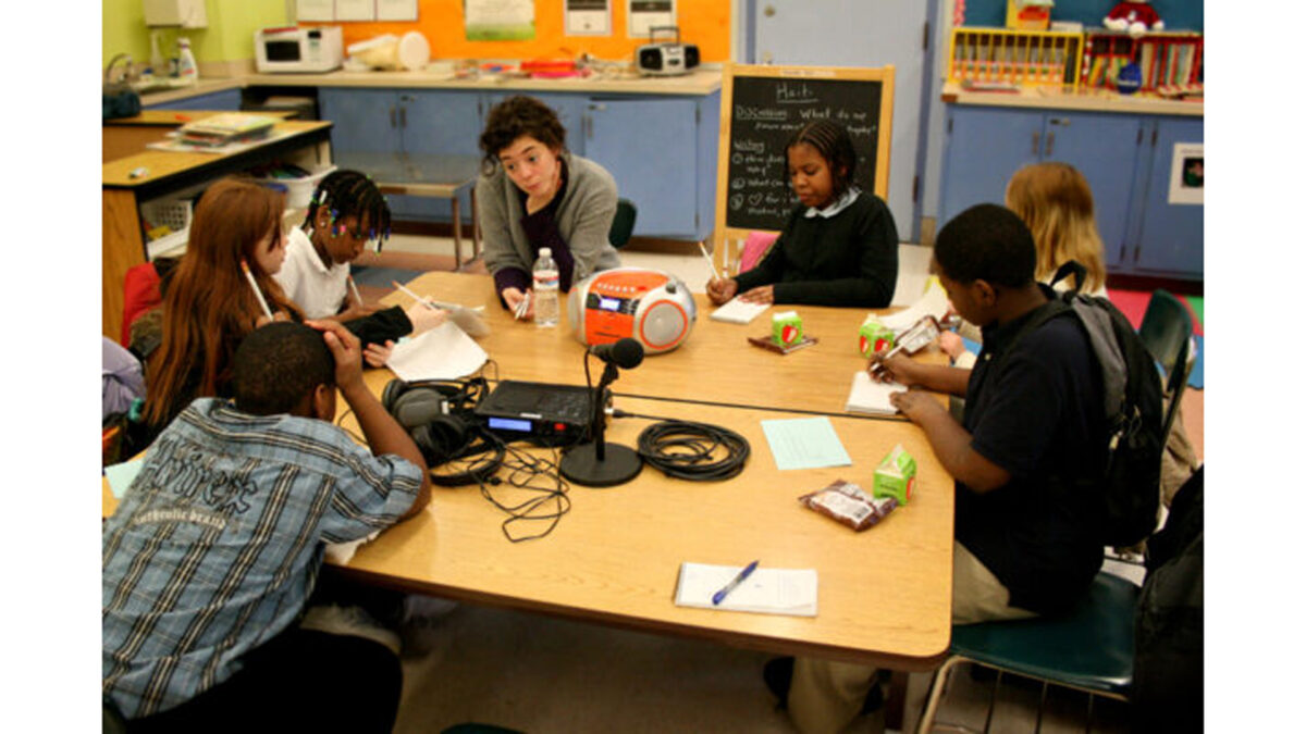 Six youth and an adult writing on pieces of paper sitting around a microphone in a classroom