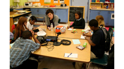 Six youth and an adult writing on pieces of paper sitting around a microphone in a classroom