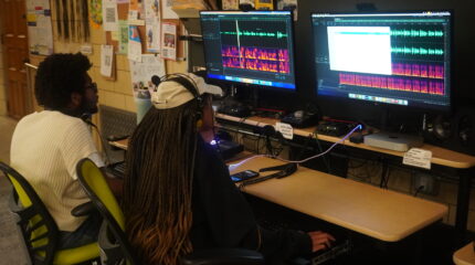 Two teens wearing headphones looking at audio editing software on computers