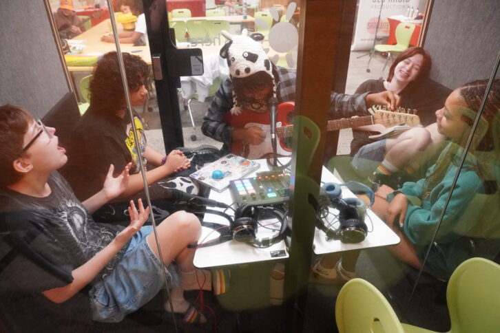 five teens seen through studio windows. one is tuning a guitar while the others smile and sing
