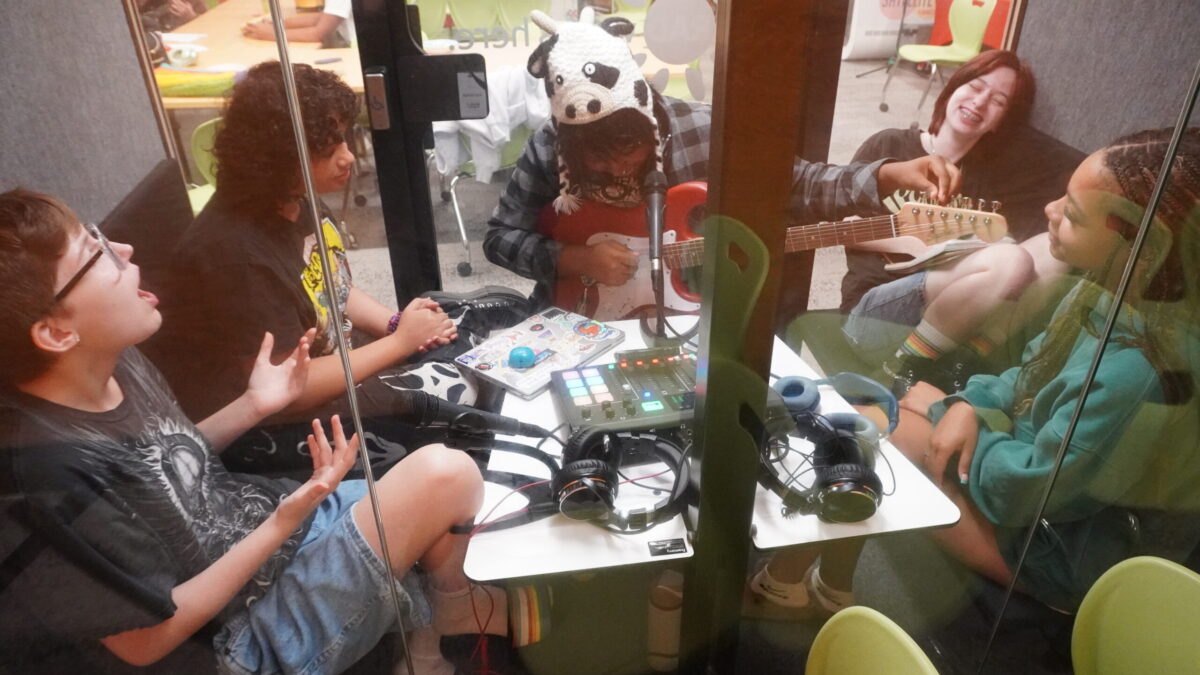 Rhyme and Reason-1 five teens seen through studio windows. one is tuning a guitar while the others smile and sing