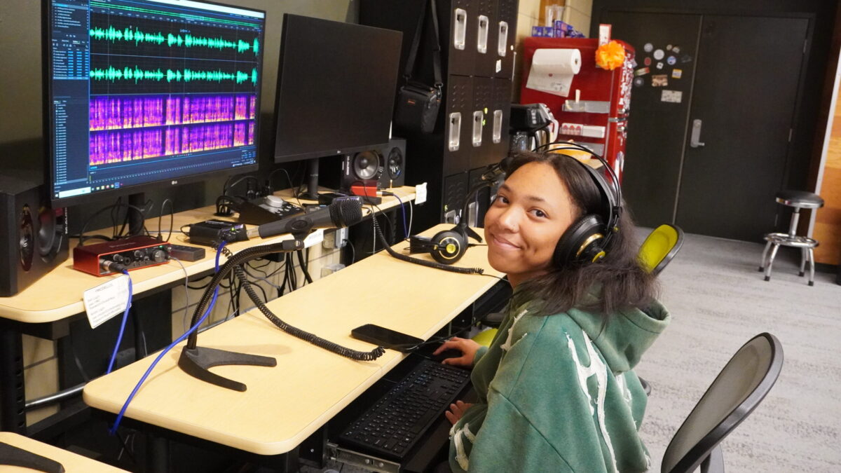A teen wearing headphones sitting at a computer displaying audio editing software