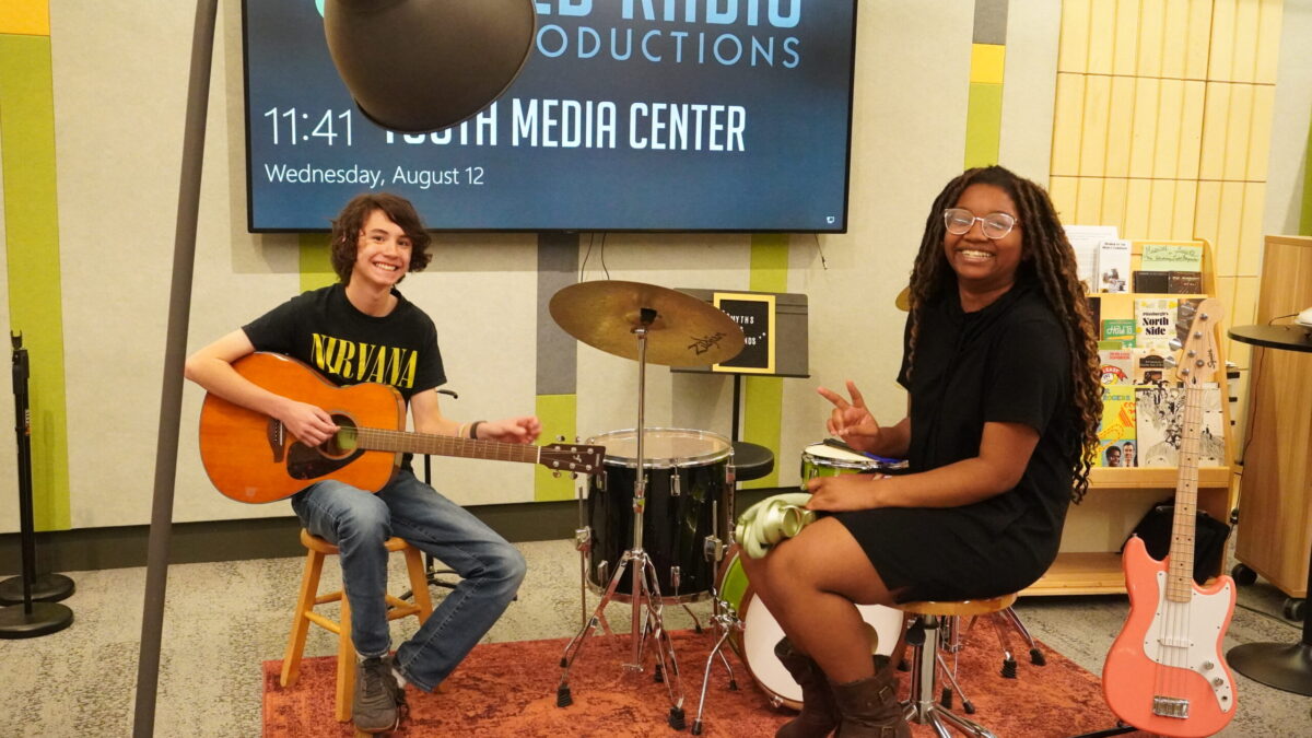 Rhyme and Reason-2 two teens smile at the camera. one holds a guitar and the other a set of headphones.