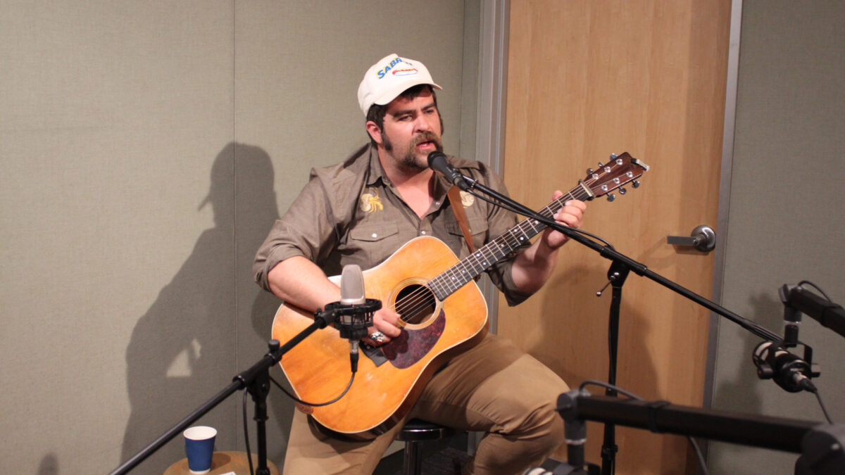 An adult playing an acoustic guitar and singing into a microphone in a radio studio