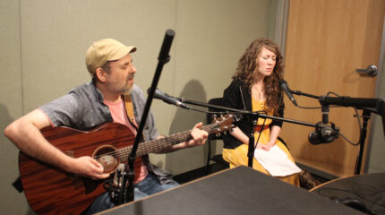 An adult playing acoustic guitar and singing into a guitar and an adult singing into a microphone in a radio studio