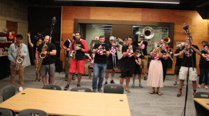 Two adults playing alto saxophones, one adult playing baritone saxophone, four adults playing trumpets, three adults and a youth playing trombones, two adults playing baritones, one adult playing tuba, three adults playing drums, and an adult dancing in the back
