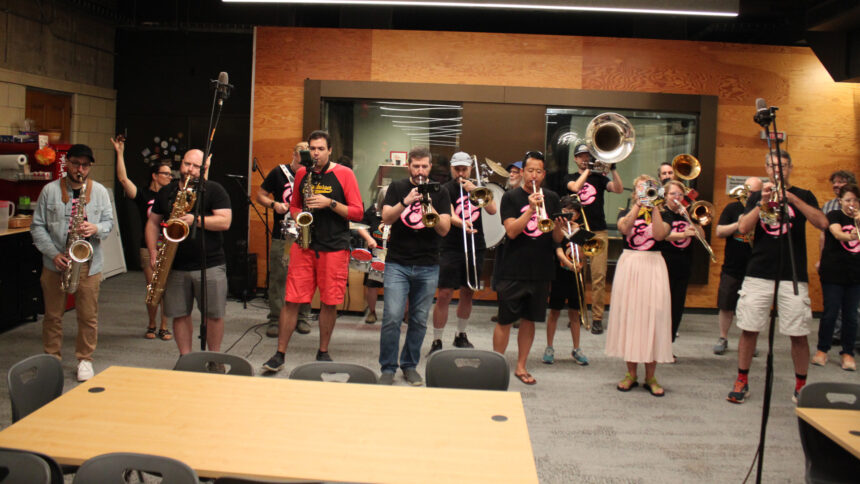 Two adults playing alto saxophones, one adult playing baritone saxophone, four adults playing trumpets, three adults and a youth playing trombones, two adults playing baritones, one adult playing tuba, three adults playing drums, and an adult dancing in the back