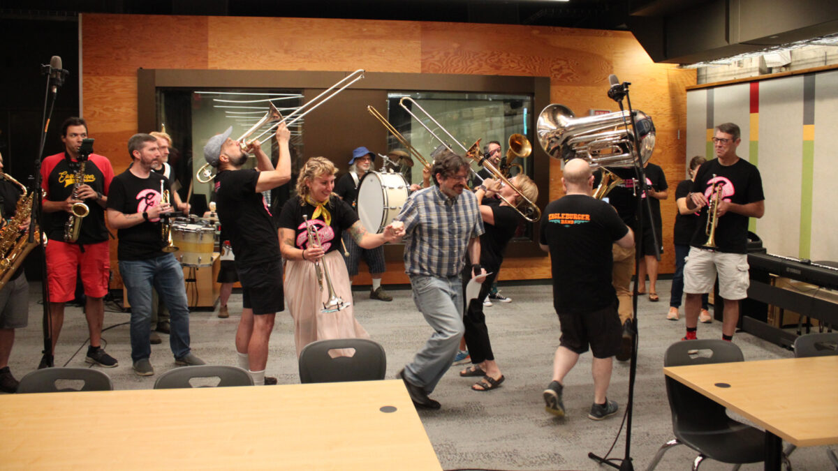 Two adults playing alto saxophones, one adult playing baritone saxophone, three adults playing trumpets, three adults and a youth playing trombones, two adults playing baritones, one adult playing tuba, three adults playing drums, and two adults dancing in the front