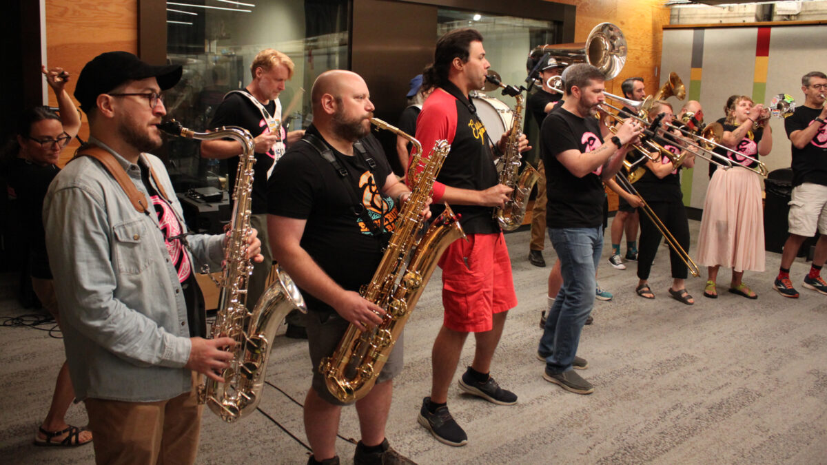 Two adults playing alto saxophones, one adult playing baritone saxophone, four adults playing trumpets, three adults and a youth playing trombones, two adults playing baritones, one adult playing tuba, three adults playing drums, and an adult dancing in the back