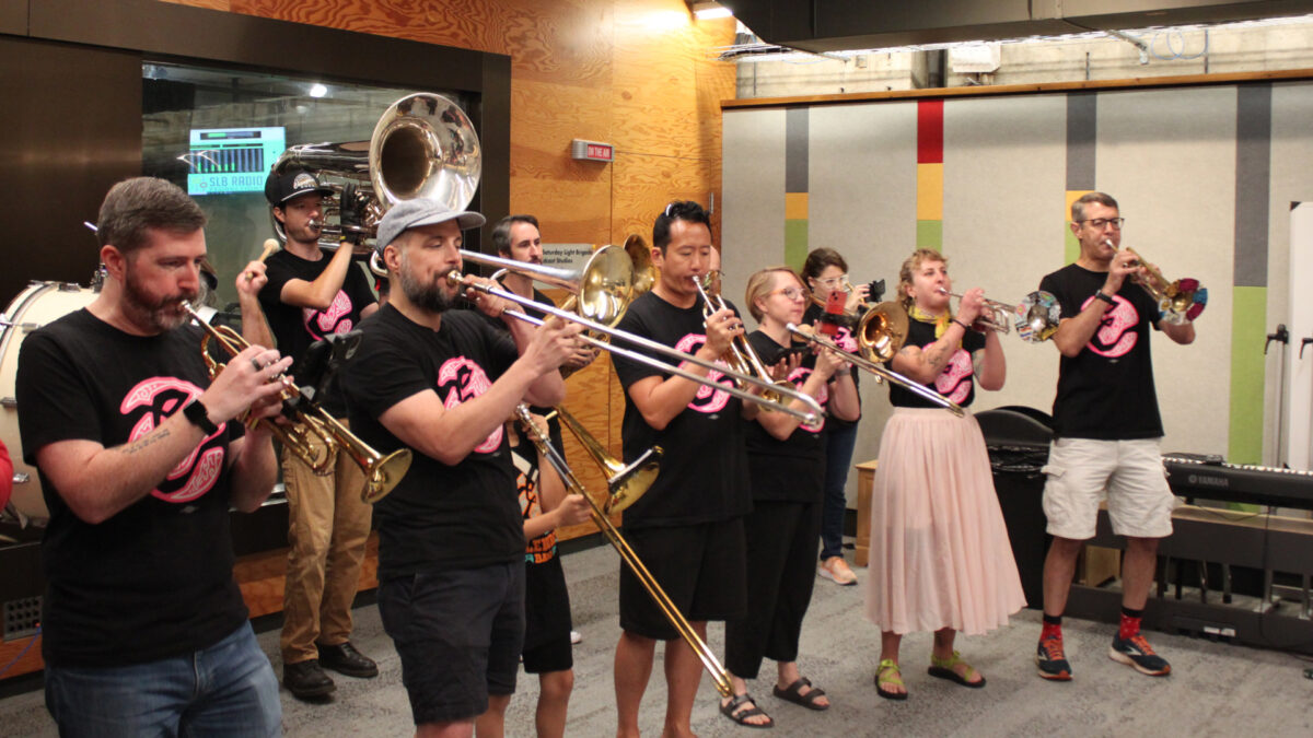 Four adults playing trumpets, three adults and a youth playing trombones, two adults playing baritones, one adult playing tuba, three adults playing drums, and an adult dancing in the back