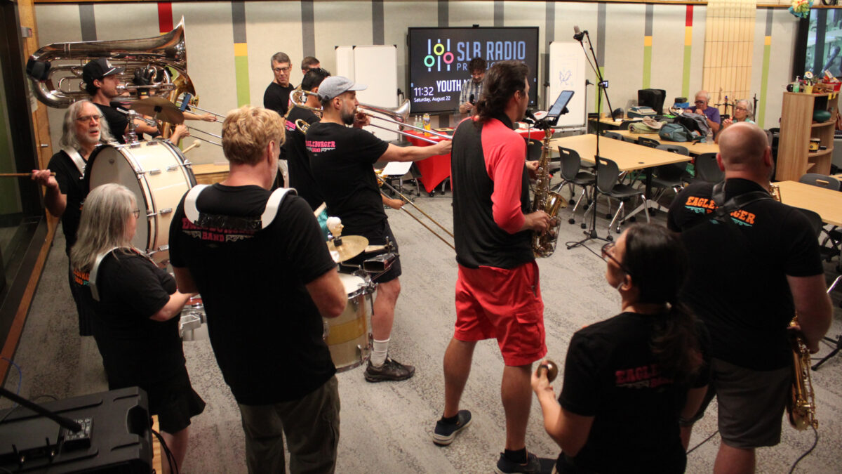 Two adults playing alto saxophones, one adult playing baritone saxophone, four adults playing trumpets, three adults and a youth playing trombones, two adults playing baritones, one adult playing tuba, three adults playing drums, and an adult dancing in the back