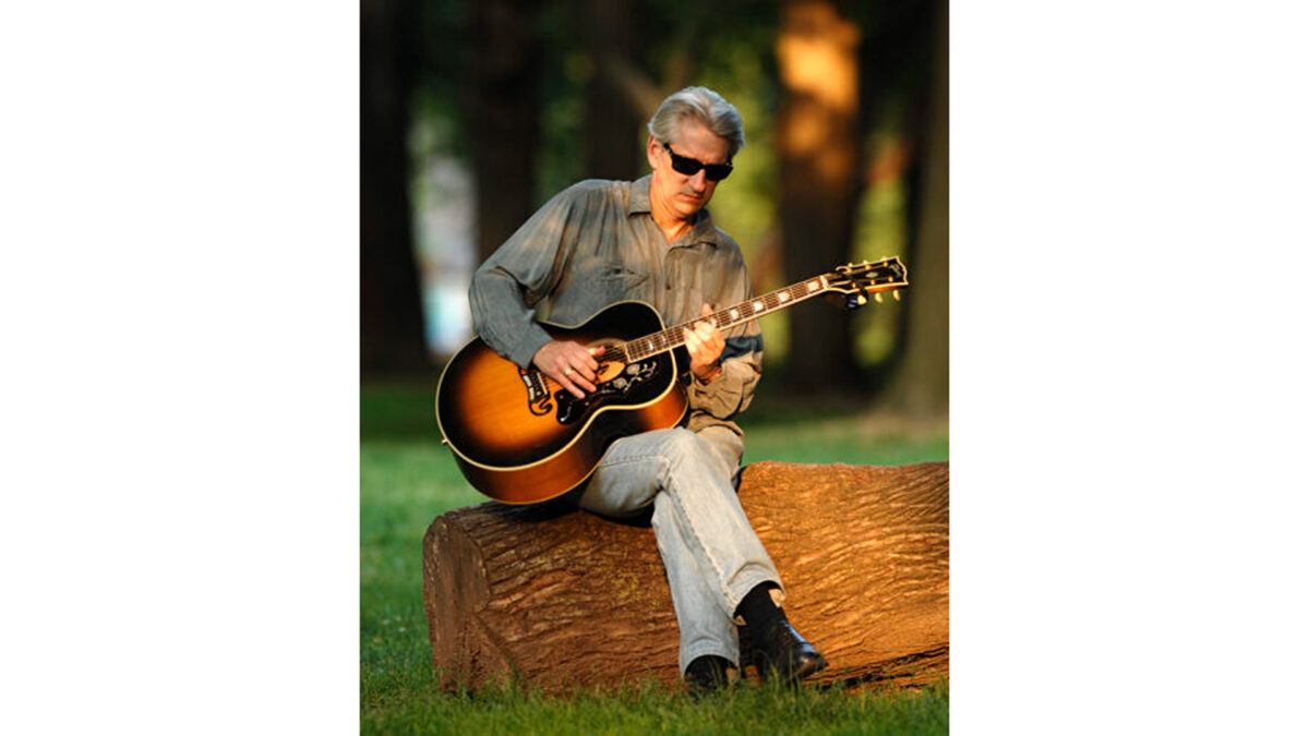 An adult wearing sunglasses sitting on a log in a forest and playing acoustic guitar