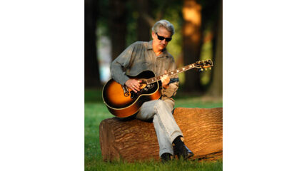 An adult wearing sunglasses sitting on a log in a forest and playing acoustic guitar