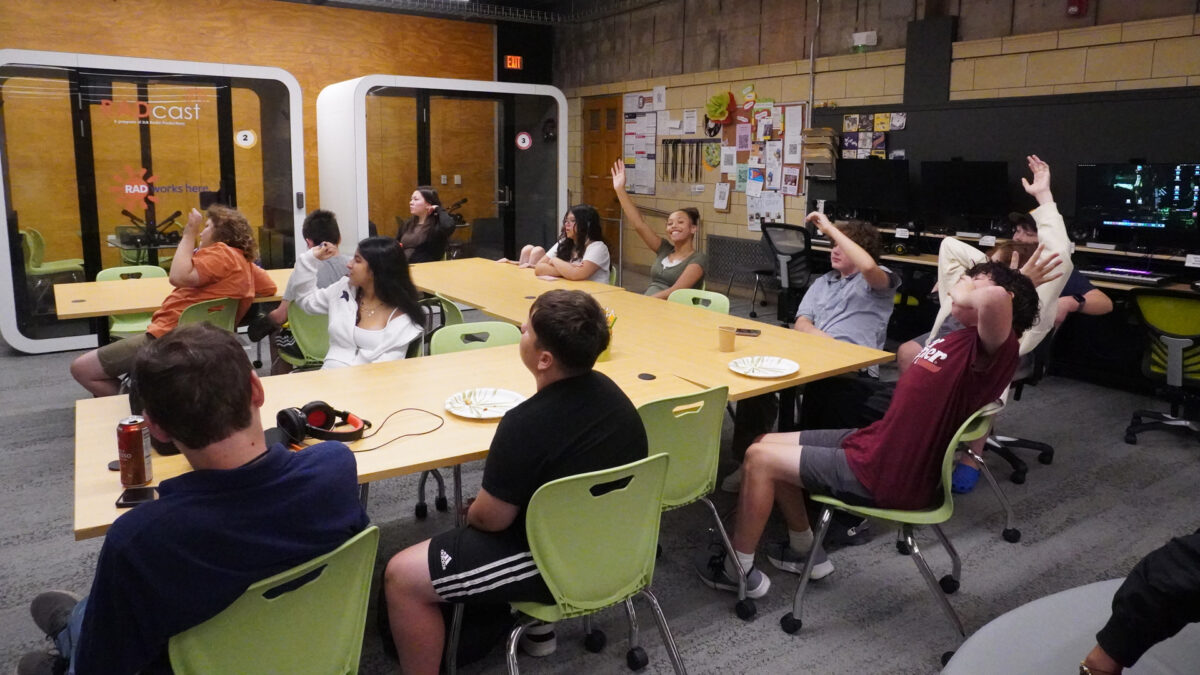 FYV-9 Ten youth and an adult sitting around tables raising an arm in the air