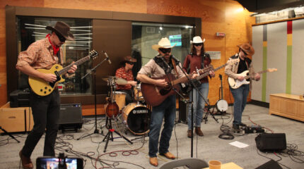Two adults playing electric guitar, an adult playing a drum set, an adult playing acoustic guitar and singing into a microphone, and an adult playing bass