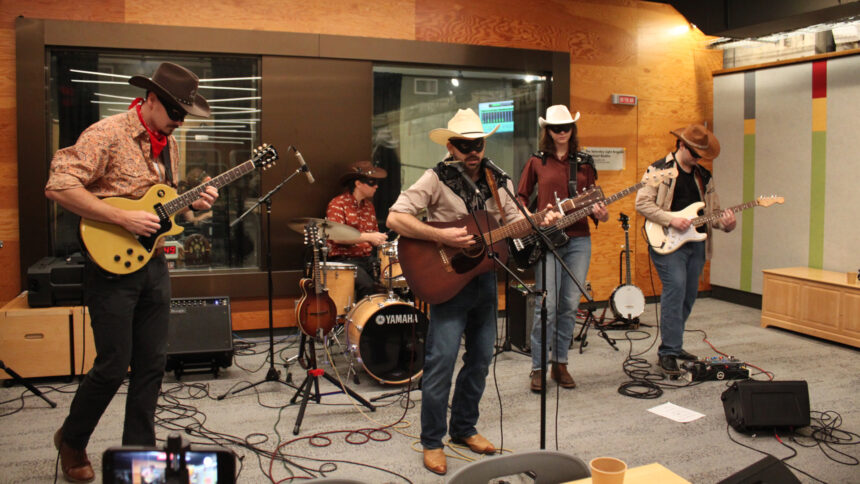 Two adults playing electric guitar, an adult playing a drum set, an adult playing acoustic guitar and singing into a microphone, and an adult playing bass