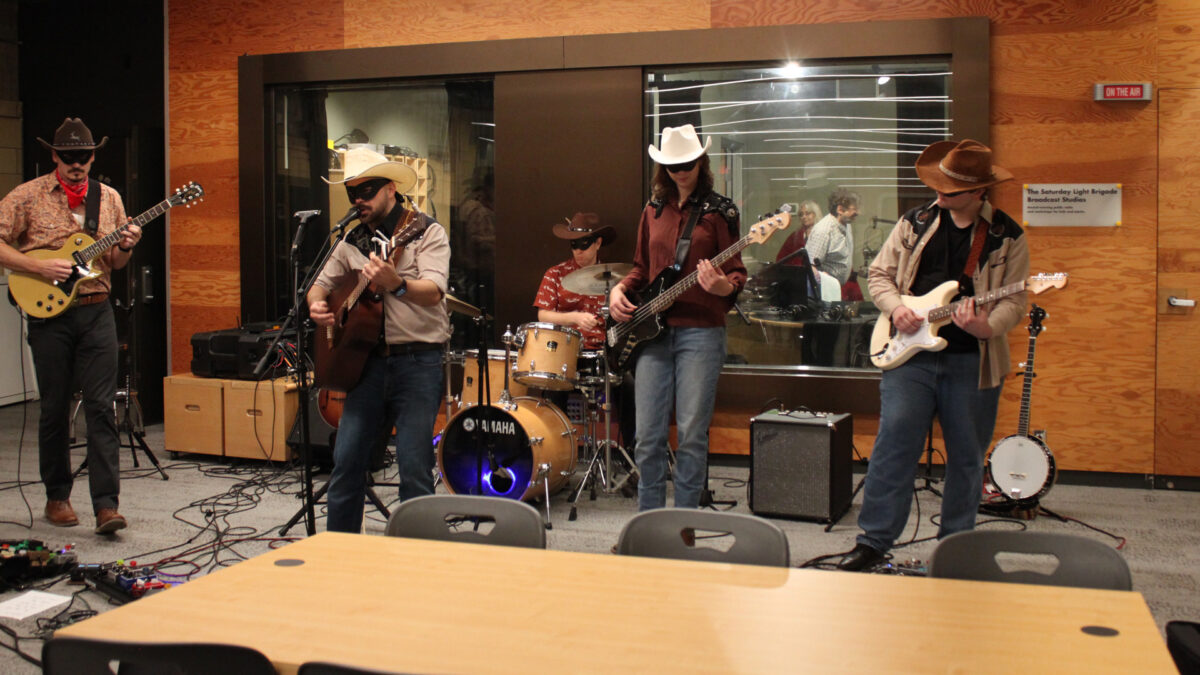 Gallop-Gallop-Gallop_2 Two adults playing electric guitar, an adult playing a drum set, an adult playing acoustic guitar and singing into a microphone, and an adult playing bass