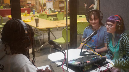 Three teens wearing headphones sitting around microphones and an audio mixer in a recording pod