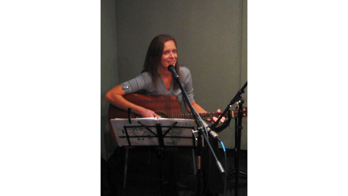 An adult playing acoustic guitar and singing into a microphone in a radio studio