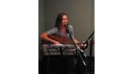 An adult playing acoustic guitar and singing into a microphone in a radio studio