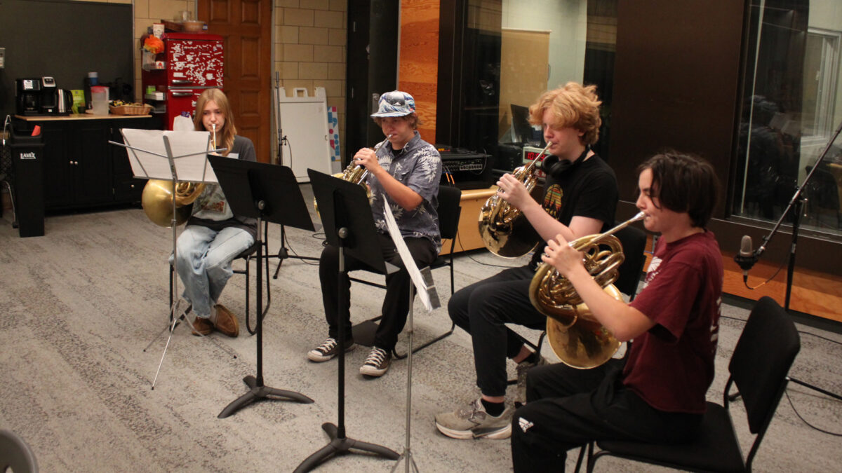 Four teens playing French horns and looking at sheet music on music stands