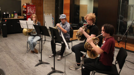Four teens playing French horns and looking at sheet music on music stands