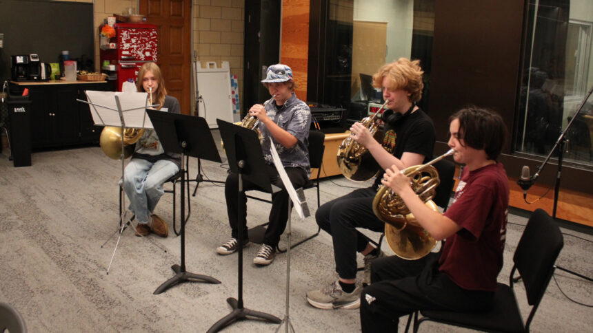 Four teens playing French horns and looking at sheet music on music stands