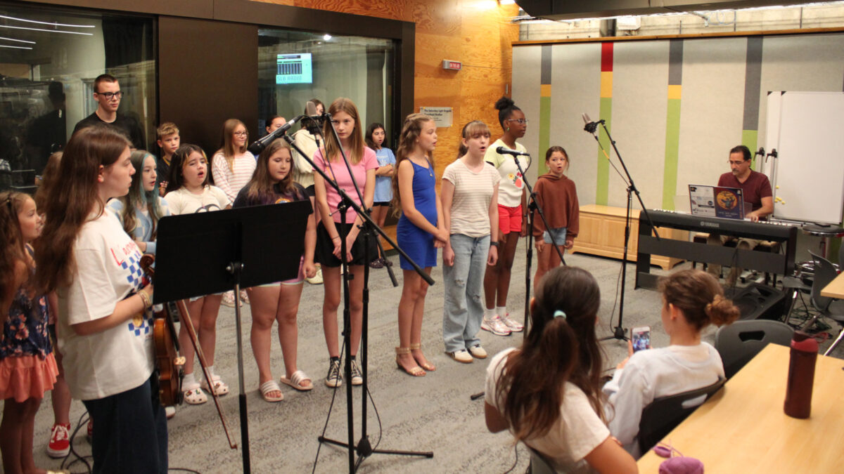 Fourteen youth singing in a choir, an adult playing a piano keyboard, and a teen holding a violin