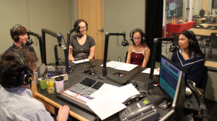 Four teens wearing headphones speaking into microphones being interviewed by an adult wearing headphones sitting behind a microphone and an audio mixer in a radio studio