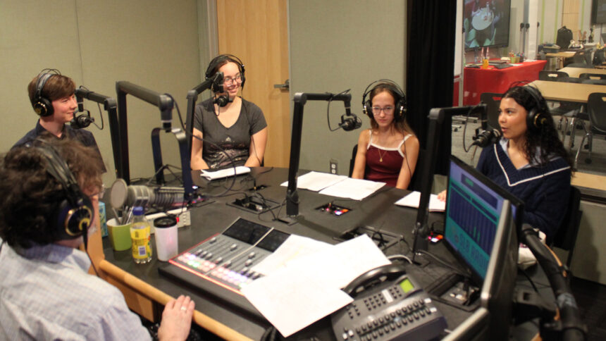 Four teens wearing headphones speaking into microphones being interviewed by an adult wearing headphones sitting behind a microphone and an audio mixer in a radio studio