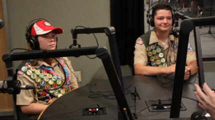 Two teens wearing headphones sitting behind microphones in a radio studio