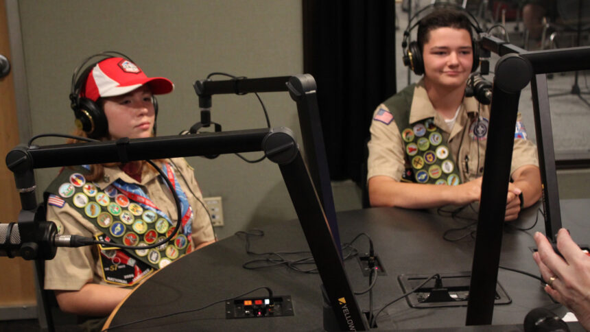 Two teens wearing headphones sitting behind microphones in a radio studio
