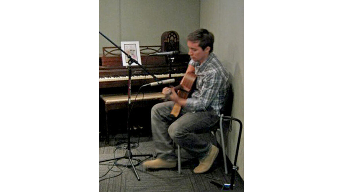 An adult playing an acoustic guitar while sitting next to a piano in a radio studio