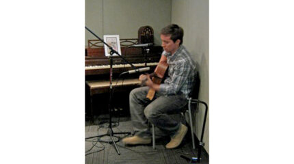 An adult playing an acoustic guitar while sitting next to a piano in a radio studio