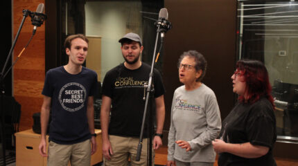 Four adults singing into microphones in front of a radio studio