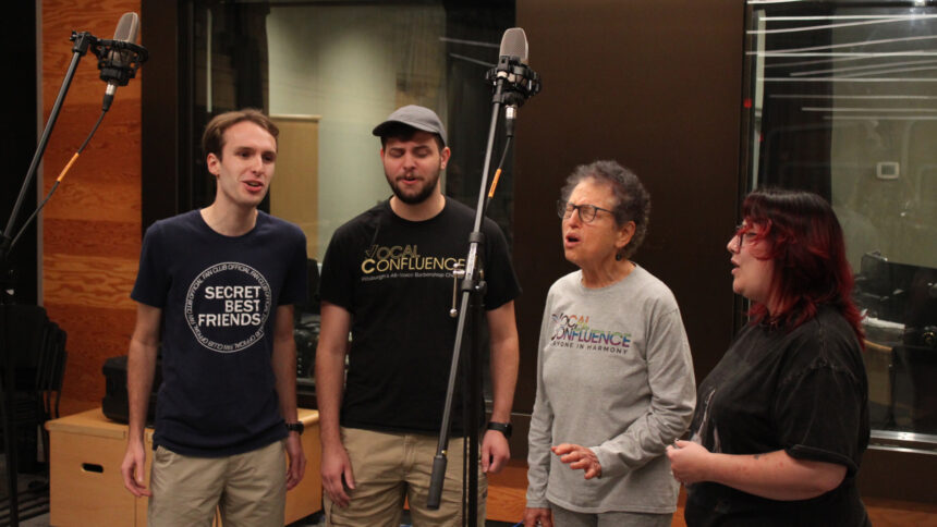 Four adults singing into microphones in front of a radio studio
