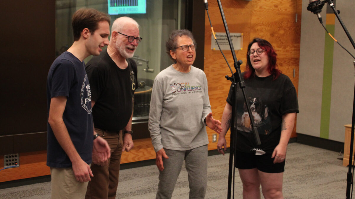 Four adults singing into microphones in front of a radio studio