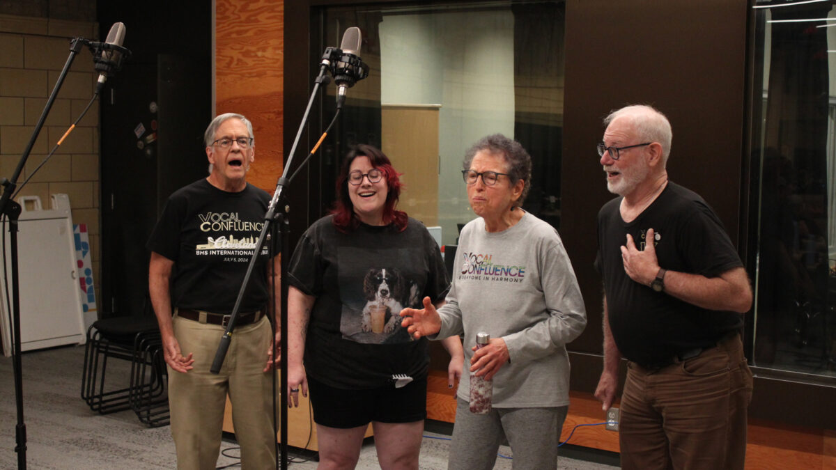 Four adults singing into microphones in front of a radio studio