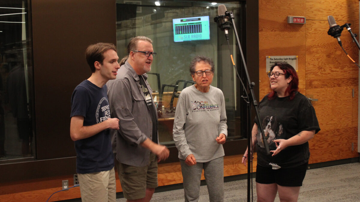 Four adults singing into microphones in front of a radio studio
