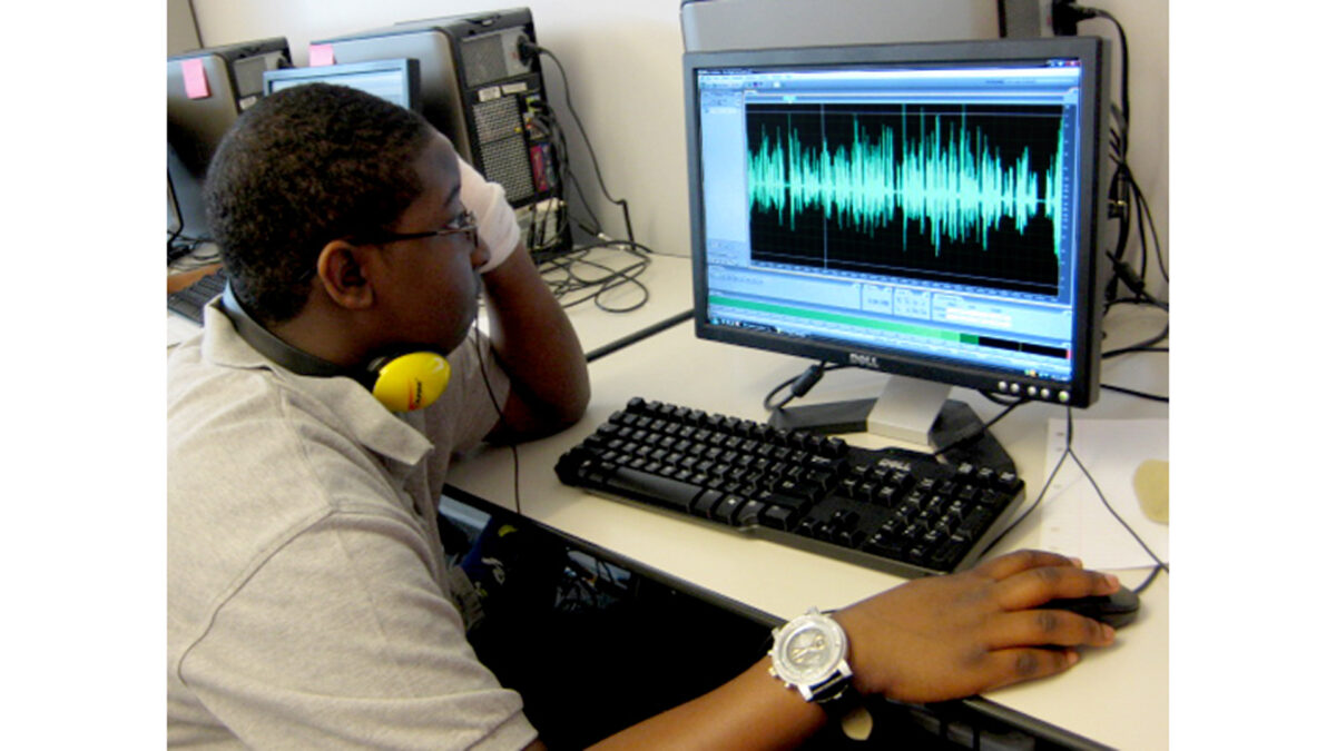 A youth wearing headphones around their neck looking at audio editing software on a computer