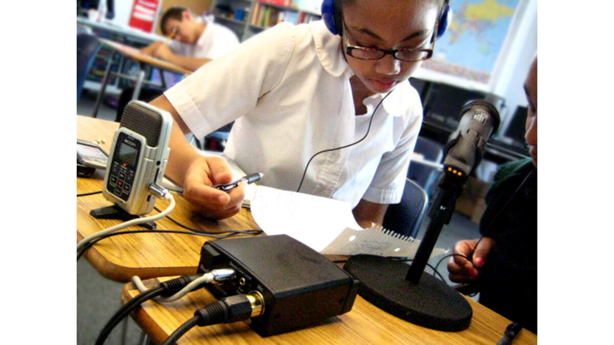 A youth wearing headphones writing on a piece of paper while sitting next to a portable recorder and a microphone